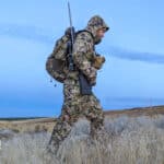 Hunter walking through dry grass with rifle on shoulder and wearing Sitka camouflage clothing
